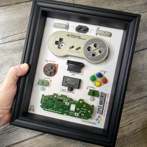 Könnte beinhalten: Eine gerahmte Darstellung von zerlegten Teilen einer Super Nintendo Entertainment System (SNES) Videospielkonsole. Die Darstellung umfasst den Controller, die Tasten, den Anschluss und das Motherboard. Der Text "SUPER NINTENDO INTRODUCED: 1990" ist auf dem weißen Hintergrund gedruckt.