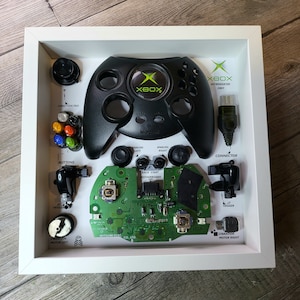 May include: A framed display of a disassembled Xbox controller, featuring the black controller, green circuit board, and various buttons and components. The frame includes labels for each part, including "Xbox Introduced 2001".