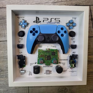 May include: A disassembled PlayStation 5 controller displayed in a white frame. The controller is blue and black and is labeled with the different parts of the controller, including the direction pad, analog sticks, buttons, and body shell. The text "PS5 INTRODUCED: 2020" is displayed at the top of the frame.