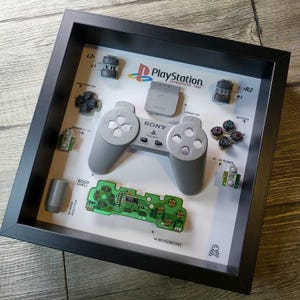 May include: A framed display of a disassembled PlayStation controller, featuring the individual components, including the body shell, buttons, and motherboard. The display also includes the PlayStation logo and the text "PlayStation. Introduced 1994".