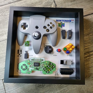 May include: A framed display of a disassembled Nintendo 64 controller. The controller is gray and has a black and green circuit board. The display also includes the text "NINTENDO 64 INTRODUCED: 1996".