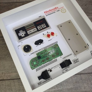 Könnte beinhalten: Ein weißer Rahmen mit weißem Hintergrund, der verschiedene Komponenten eines Nintendo Entertainment Systems zeigt. Die Komponenten umfassen den Controller, das Motherboard und den Anschluss. Der Text "Nintendo Entertainment System Introduced: 1983" befindet sich oben im Rahmen.