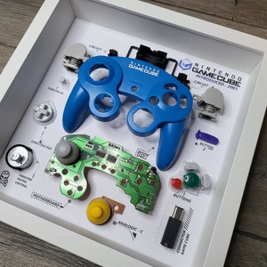 Puede incluir: Un mando de Nintendo GameCube desmontado que se muestra en un marco blanco. El mando es azul y se ha separado en sus componentes individuales, incluyendo la placa de circuito, los botones y la palanca analógica. El marco incluye etiquetas que identifican cada parte.