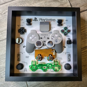 May include: A framed display of a disassembled PlayStation controller. The controller is shown in its component parts, including the buttons, analog sticks, circuit board, and vibration motors. The frame is black and the background is white with black text.