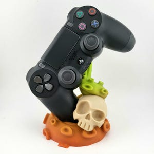 Può includere: Un controller PlayStation 4 nero è posizionato su un supporto stampato in 3D. Il supporto ha la forma di un teschio con una pianta verde che cresce dalla cima. La base del supporto è arancione.