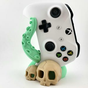 Può includere: Un controller Xbox bianco con un'impugnatura verde a forma di polpo è esposto su un supporto realizzato con due statuette di teschi beige. Il supporto è su una base verde.