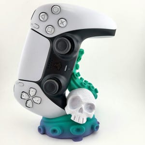 Peut inclure: Une manette de jeu PlayStation 5 blanche est placée sur un support imprimé en 3D. Le support est en forme de pieuvre avec un crâne blanc au centre. La pieuvre est verte et bleue.