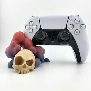 Puede incluir: Un mando de PlayStation 5 blanco y negro descansa sobre un soporte en forma de calavera impreso en 3D. El soporte está hecho de resina multicolor con tonos rojos, morados y azules.