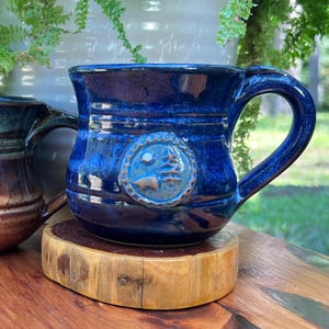 May include: A handmade, cobalt blue ceramic mug with a large handle. The mug features a circular emblem with a mountain and tree design. The mug is sitting on a wooden coaster. The mug is a unique piece of drinkware.
