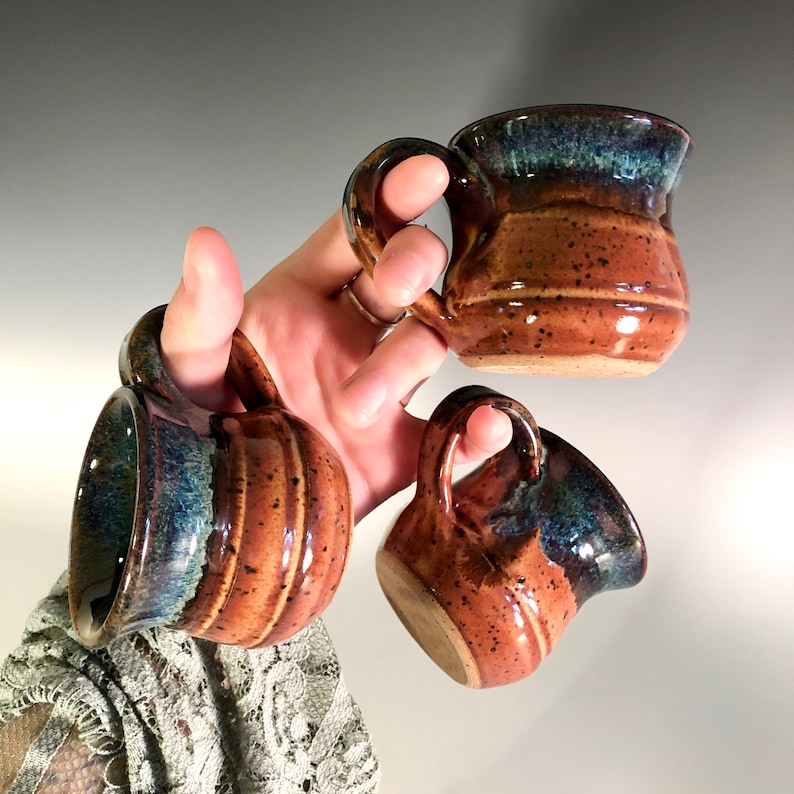 May include: Three handmade ceramic mugs with handles. The mugs have a speckled brown base with a blue and green glazed rim. The mugs are held in a hand against a neutral background. The mugs are a unique addition to any kitchen.