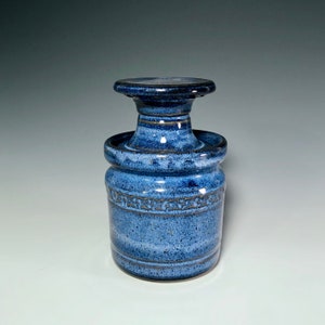 May include: A handcrafted blue ceramic butter dish with a lid. The dish has a cylindrical base with a decorative band and a flared top. The lid is a flat disc shape. The glaze has a speckled appearance.