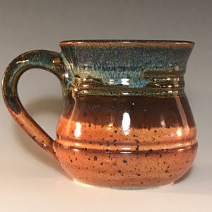 May include: A handmade ceramic mug with a rounded body and a large handle. The mug features a gradient of colors, transitioning from a speckled orange base to a dark brown middle and a blue-green rim. The mug is approximately 4 inches tall.