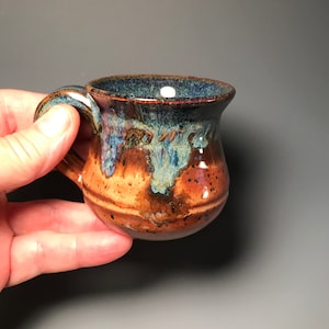 May include: A small, handmade ceramic mug with a rounded body and a handle. The mug features a blend of brown, blue, and tan colors with a speckled texture. The interior is a deep blue, and the exterior has a glossy finish.