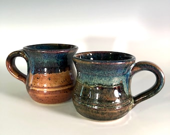 May include: Two handmade ceramic mugs with a rustic design. The mugs feature a dark blue and brown glaze, with a rounded handle. The mugs are approximately 4 inches tall and 3 inches in diameter.