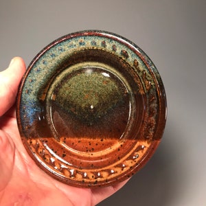 May include: A handmade ceramic dish with a circular shape. The dish features a gradient of colors, including brown, green, and blue. The rim has a textured design. The dish is likely used for serving or display.