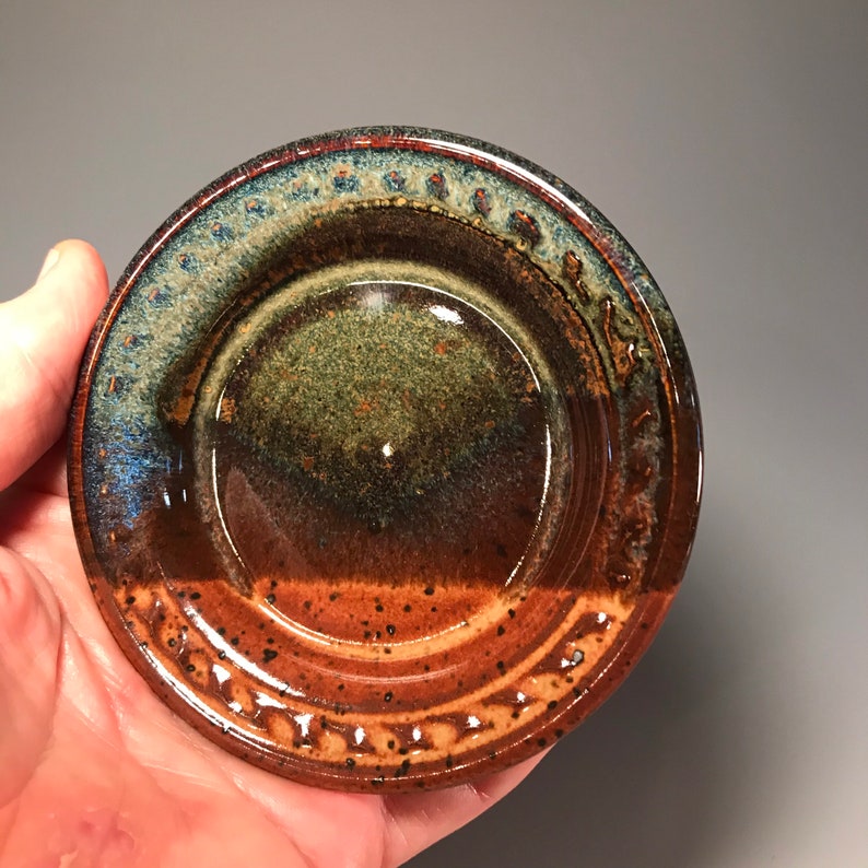 May include: A handmade ceramic dish with a circular shape. The dish features a gradient of colors, including brown, green, and blue. The rim has a textured design. The dish is likely used for serving or display.