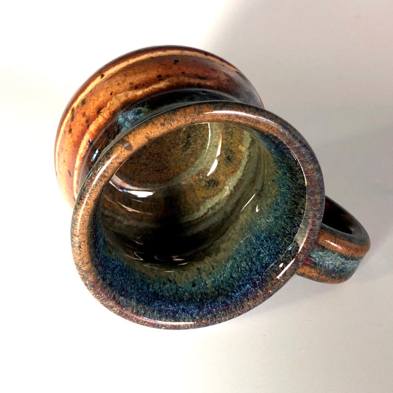 May include: A handmade ceramic mug with a unique glaze. The mug features a brown and blue color scheme with a glossy finish. The handle is small and rounded. The interior of the mug has a dark blue and green color.