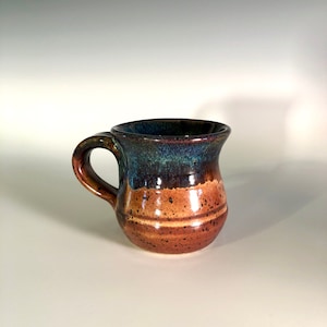 May include: A handmade ceramic mug with a rounded body and a curved handle. The mug features a color gradient, transitioning from a dark blue-green rim to a brown and tan speckled base. The mug is approximately 4 inches tall.