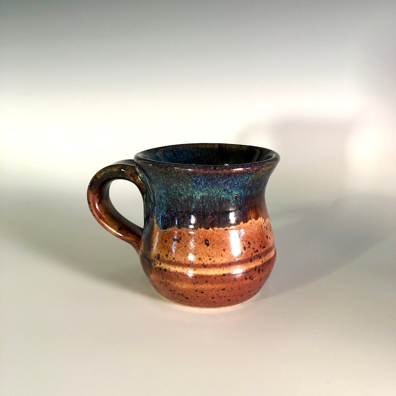 May include: A handmade ceramic mug with a rounded body and a curved handle. The mug features a color gradient, transitioning from a dark blue-green rim to a brown and tan speckled base. The mug is approximately 4 inches tall.