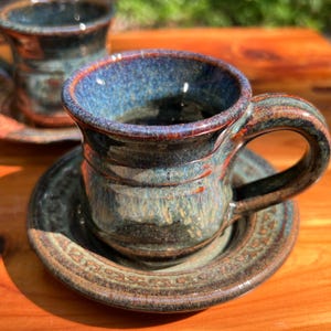 May include: Handmade ceramic cup and saucer set. The cup has a rounded shape with a handle and a blue and brown glaze. The saucer is a matching color and design. The set is on a wooden surface.