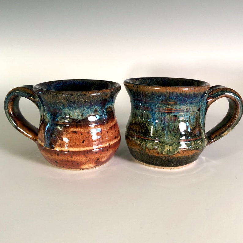 May include: Two handmade ceramic mugs with a unique, textured glaze. One mug features brown and tan horizontal stripes with a blue rim. The other mug has a green and blue glaze. Both mugs have a rounded shape and a sturdy handle.