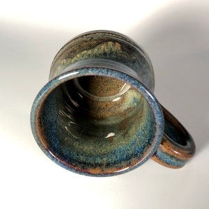 May include: A handmade ceramic mug with a unique glaze. The mug features a blend of blue, brown, and green hues, creating a marbled effect. It has a sturdy handle and a wide opening, perfect for coffee or tea. The mug is approximately 4 inches tall.