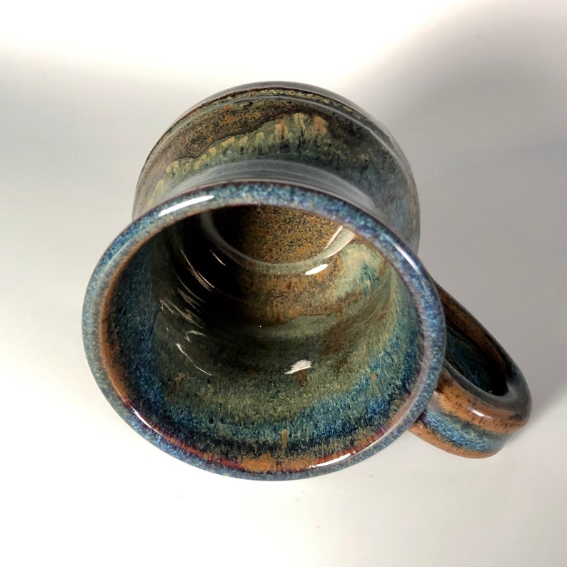 May include: A handmade ceramic mug with a unique glaze. The mug features a blend of blue, brown, and green hues, creating a marbled effect. It has a sturdy handle and a wide opening, perfect for coffee or tea. The mug is approximately 4 inches tall.