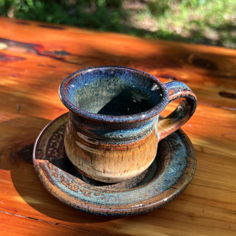 May include: Handmade ceramic espresso cup and saucer set. The cup features a dark blue rim that fades into a brown and tan body. The saucer has a matching blue and brown glaze. The set is perfect for enjoying a morning espresso.