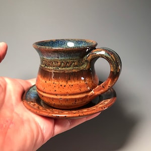 May include: A handmade ceramic espresso cup and saucer set. The cup features a rounded body with a handle and a dark blue rim. The exterior transitions from orange to brown. The saucer is a matching color. The set is held in a hand.