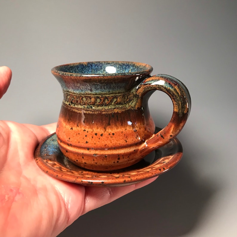May include: A handmade ceramic espresso cup and saucer set. The cup features a rounded body with a handle and a dark blue rim. The exterior transitions from orange to brown. The saucer is a matching color. The set is held in a hand.