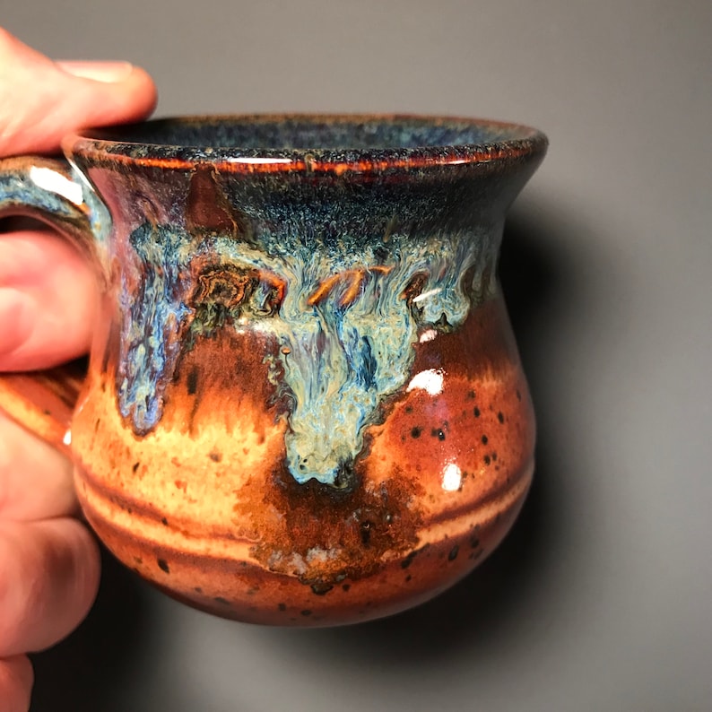 May include: A handmade ceramic mug with a unique glaze. The mug features a brown base with a gradient of orange and blue hues. The interior and rim are dark blue. The handle is a matching brown color.