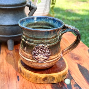 May include: A handcrafted ceramic mug with a campfire emblem. The mug features a blue interior, a brown exterior with green and tan accents, and a sturdy handle. It sits on a wooden coaster, perfect for enjoying hot beverages.