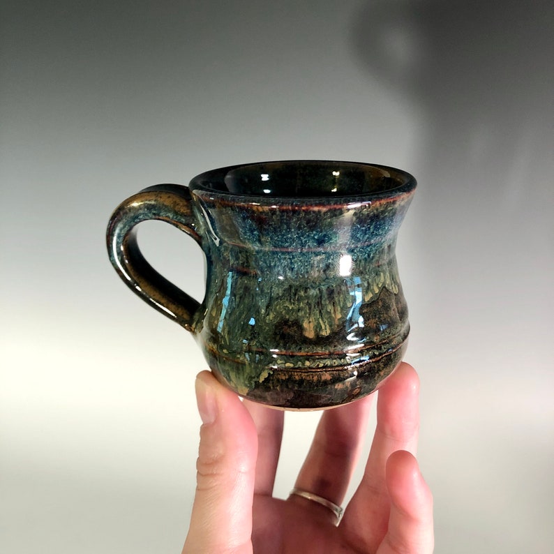 May include: A handmade ceramic mug with a dark blue, green, and brown glaze. The mug has a rounded body, a sturdy handle, and a glossy finish. The mug is approximately 3 inches tall.