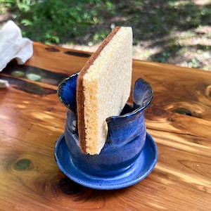 May include: A handmade, cobalt blue ceramic sponge holder with a matching saucer. The holder has a unique, organic shape with a textured surface. A beige and brown sponge is placed inside the holder. The holder is sitting on a wooden surface.