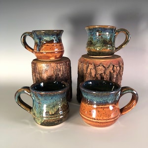 May include: Four handmade ceramic mugs with a rustic design. The mugs feature a dark blue glaze on the rim, transitioning to brown and orange tones. Each mug has a handle and is displayed on a small wooden stand.