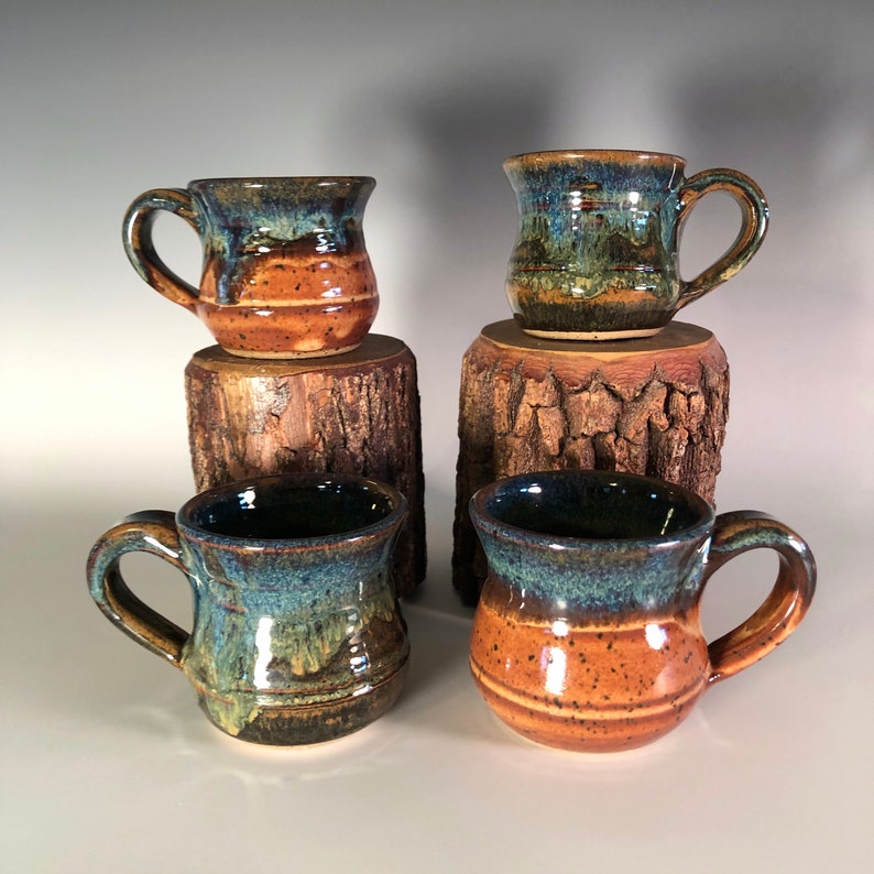 May include: Four handmade ceramic mugs with a rustic design. The mugs feature a dark blue glaze on the rim, transitioning to brown and orange tones. Each mug has a handle and is displayed on a small wooden stand.