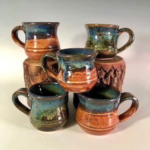 May include: A collection of five handmade ceramic mugs. Each mug features a unique blend of colors, including brown, blue, and green, with a glossy finish. The mugs are displayed on wooden blocks, showcasing their rustic charm and artisanal quality.
