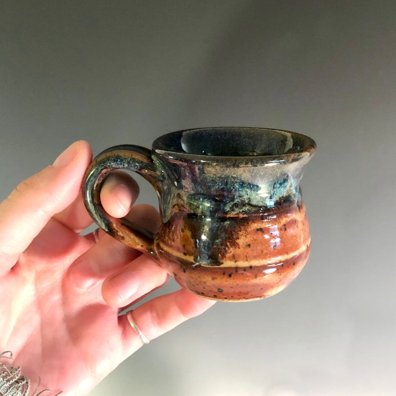 May include: A small, handmade ceramic mug with a rounded handle. The mug features a speckled brown base with horizontal bands and a dark blue and green rim. The interior is dark blue. The mug is held in a hand.