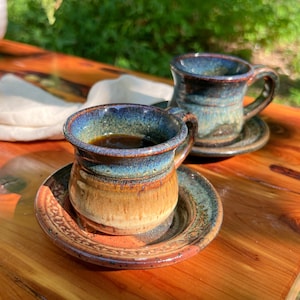 May include: Two handmade ceramic coffee mugs with saucers. The mugs have a blue and brown glaze, and are filled with dark coffee. The saucers have a matching design. The mugs are on a wooden surface.