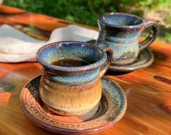 May include: Two handmade ceramic coffee mugs with saucers. The mugs have a blue and brown glaze, and are filled with dark coffee. The saucers have a matching design. The mugs are on a wooden surface.