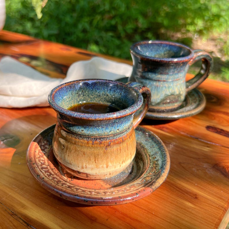 May include: Two handmade ceramic coffee mugs with saucers. The mugs have a blue and brown glaze, and are filled with dark coffee. The saucers have a matching design. The mugs are on a wooden surface.