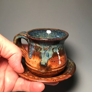 May include: Handmade ceramic espresso cup and saucer set. The cup features a dark blue rim, with brown and blue glaze. The saucer has a matching brown glaze. The cup is approximately 2.5 inches tall and 2 inches wide.