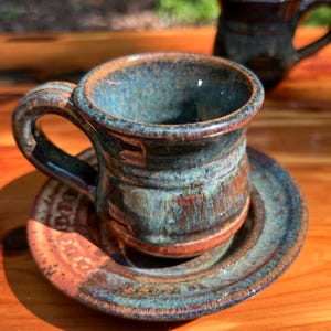 May include: A handmade ceramic mug and saucer set. The mug features a rounded shape with a handle, and the saucer is circular. The set is glazed in shades of blue, brown, and orange, with a textured finish. The mug is approximately 3 inches tall.