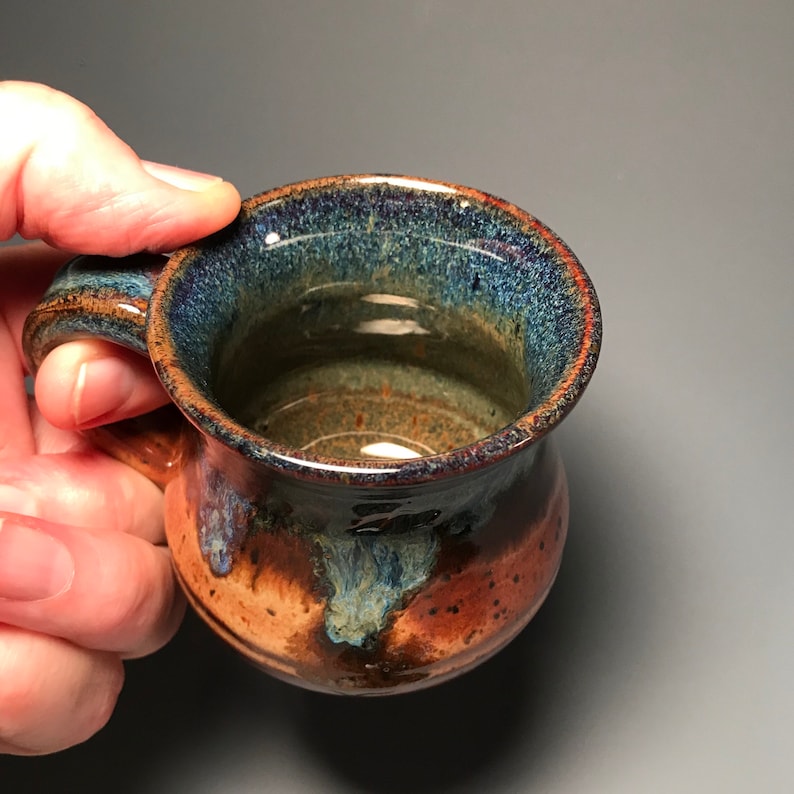 May include: A handmade ceramic mug with a rounded body and a handle. The mug features a blend of colors, including brown, blue, and hints of red. The interior is a lighter brown, and the rim has a darker, textured appearance. The mug is held by a hand.