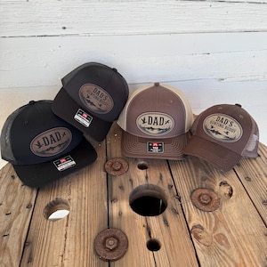 May include: A collection of baseball caps in various shades of brown and black, featuring the text "DAD" or "DAD'S HUNTING BUDDY" on a leather patch. The caps have a mesh back and a curved brim.