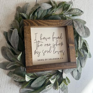May include: A small, square wooden sign with the quote "I have found the one whom my soul loves. Song of Solomon 3:4" in black script. The sign is framed in dark brown wood and rests on a bed of green and gray leaves.