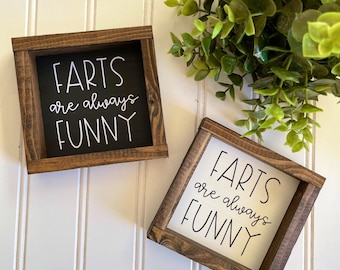 May include: Two wooden signs with a black and white background. The signs have a brown wooden frame and say "Farts are always funny" in white cursive writing.