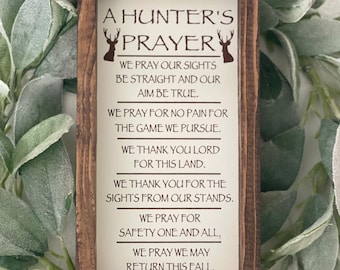 May include: A wooden sign with a hunter's prayer. The sign is white with brown text and two deer silhouettes. The text reads: "A Hunter's Prayer. We pray our sights be straight and our aim be true. We pray for no pain for the game we pursue. We thank you Lord for this land. We thank you for the sights from our stands. We pray for safety one and all. We pray we may return this fall."