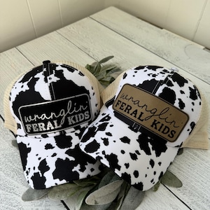May include: Two trucker hats with a black and white cow print design. Each hat has a rectangular patch with the words "wranglin' FERAL KIDS" in black and white or brown lettering.