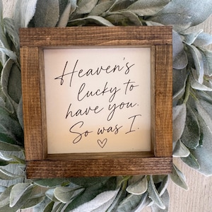May include: A square wooden sign with a dark brown stained frame and a white center panel. The text in black cursive script reads: Heaven's lucky to have you. So was I. A small black heart icon is centered below the text on the white background.